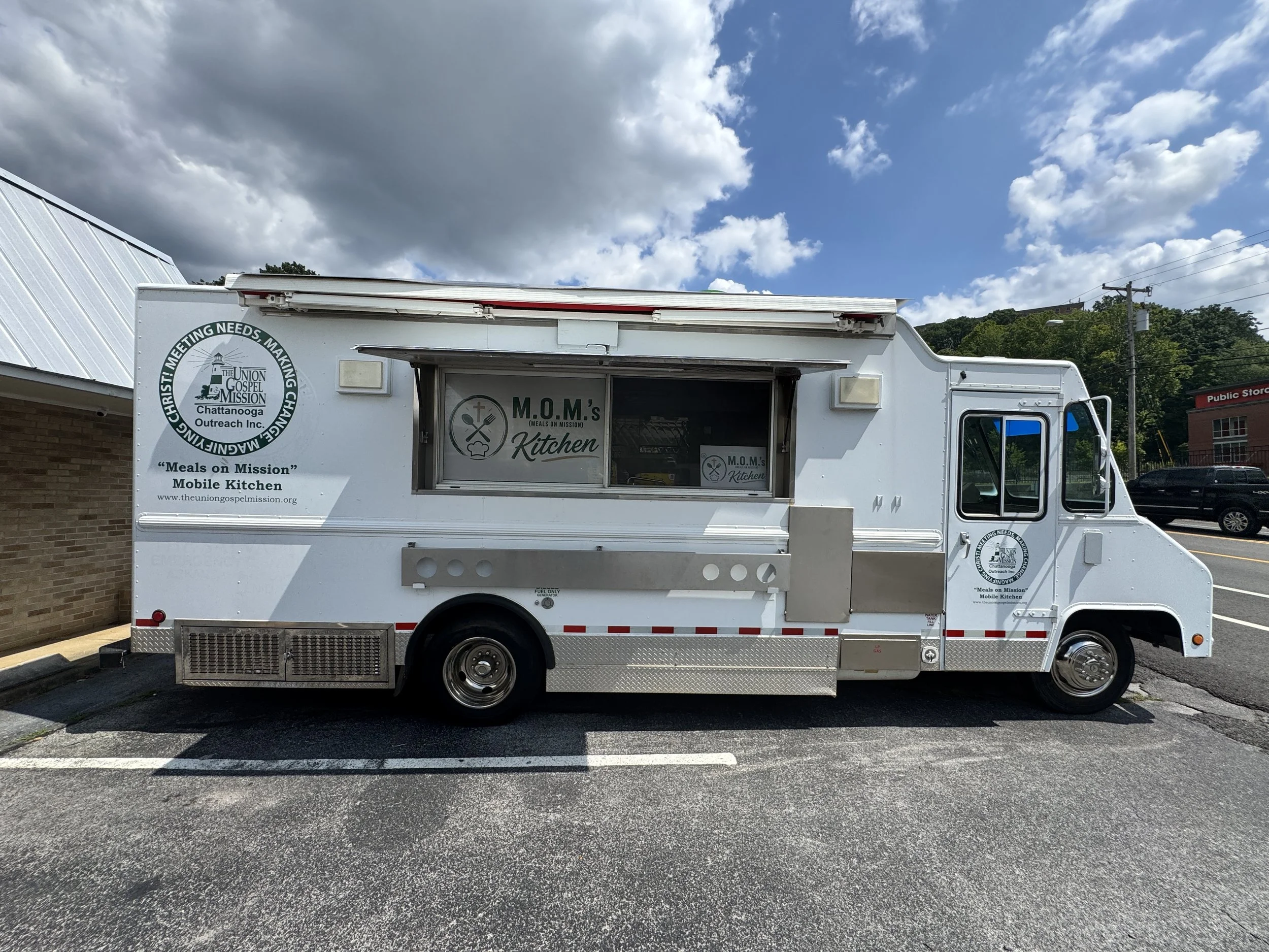 A white mobile kitchen truck with logos and text on the side, parked in a parking lot under a partly cloudy sky. The truck has a window and a small serving counter.