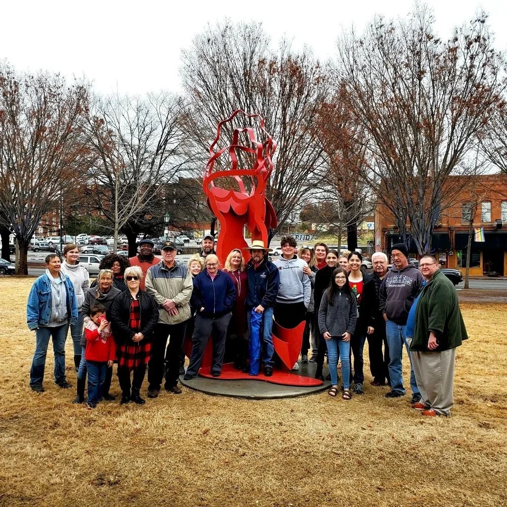Group of diverse people gathered outdoors around a red metal sculpture of the shape of a human fist with a heart in place of the palm, with leafless trees and buildings in the background on a cloudy day.