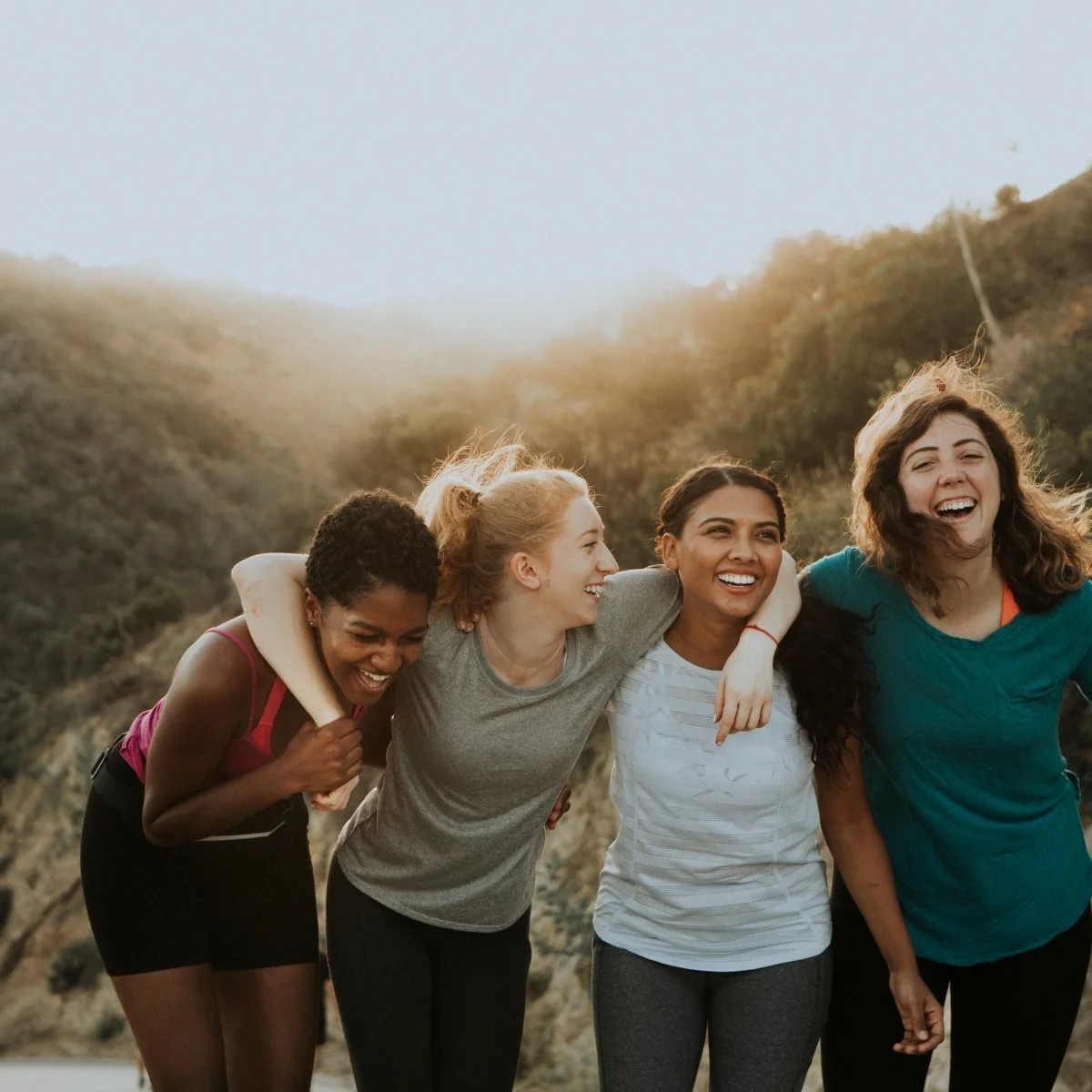 Four women outdoors, laughing and embracing on a sunny day, with a natural landscape in the background.