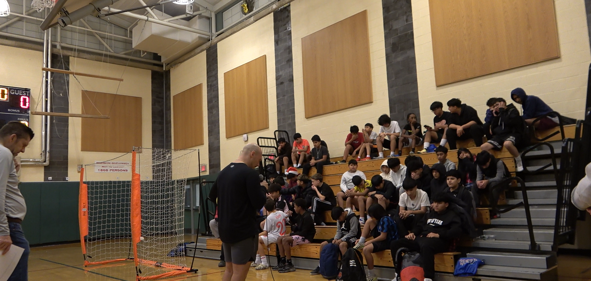 Group of students sitting on wooden bleachers in a gymnasium, with a coach standing nearby, a small soccer goal, and a scoreboard in the background