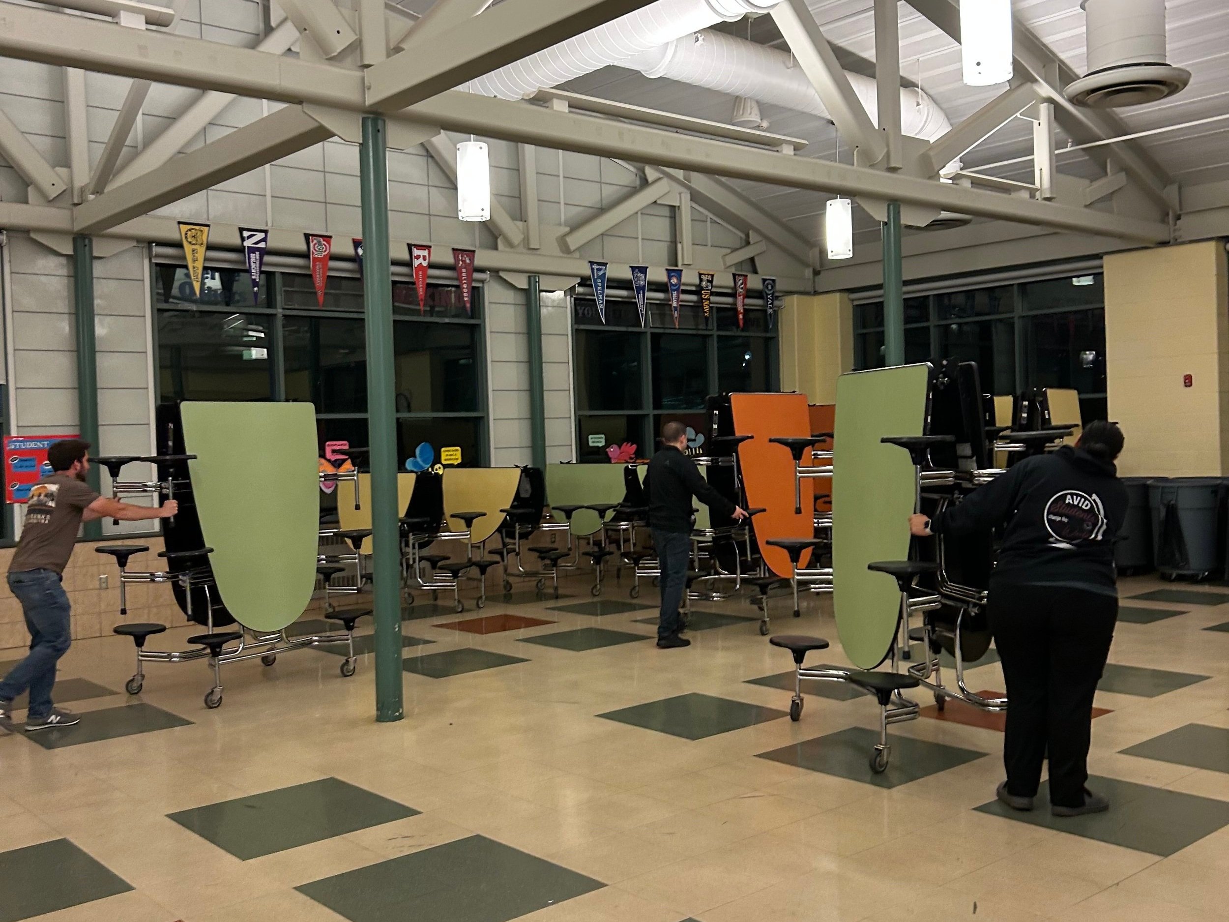 Three people arranging stacked chairs and tables in a school cafeteria, with colorful banners hanging from the ceiling.
