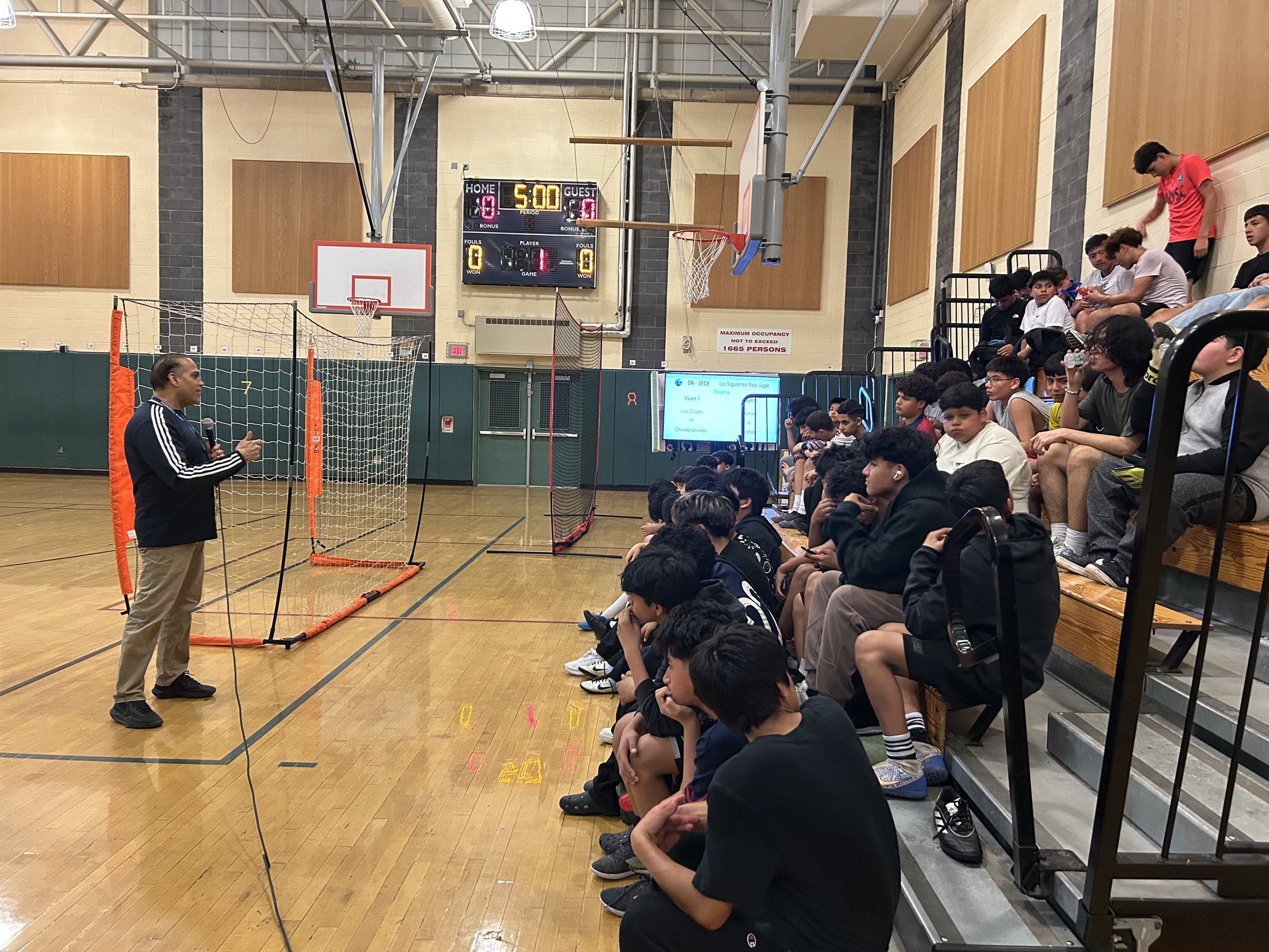 The school superintendent in a navy jacket speaks to a group of students seated on gym bleachers inside a gymnasium with a basketball hoop overhead, a scoreboard showing 5:00, and soccer goals on the floor.