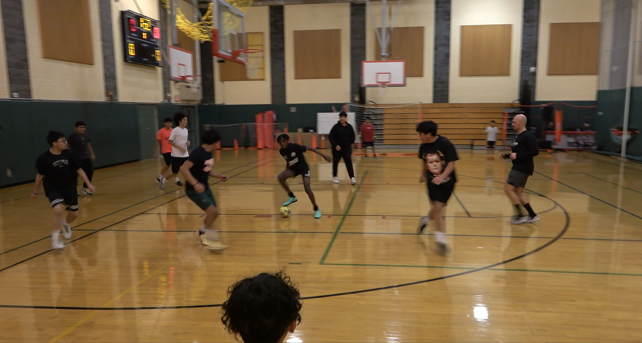 Children playing indoor soccer on a gymnasium court with basketball hoops and scoreboard.