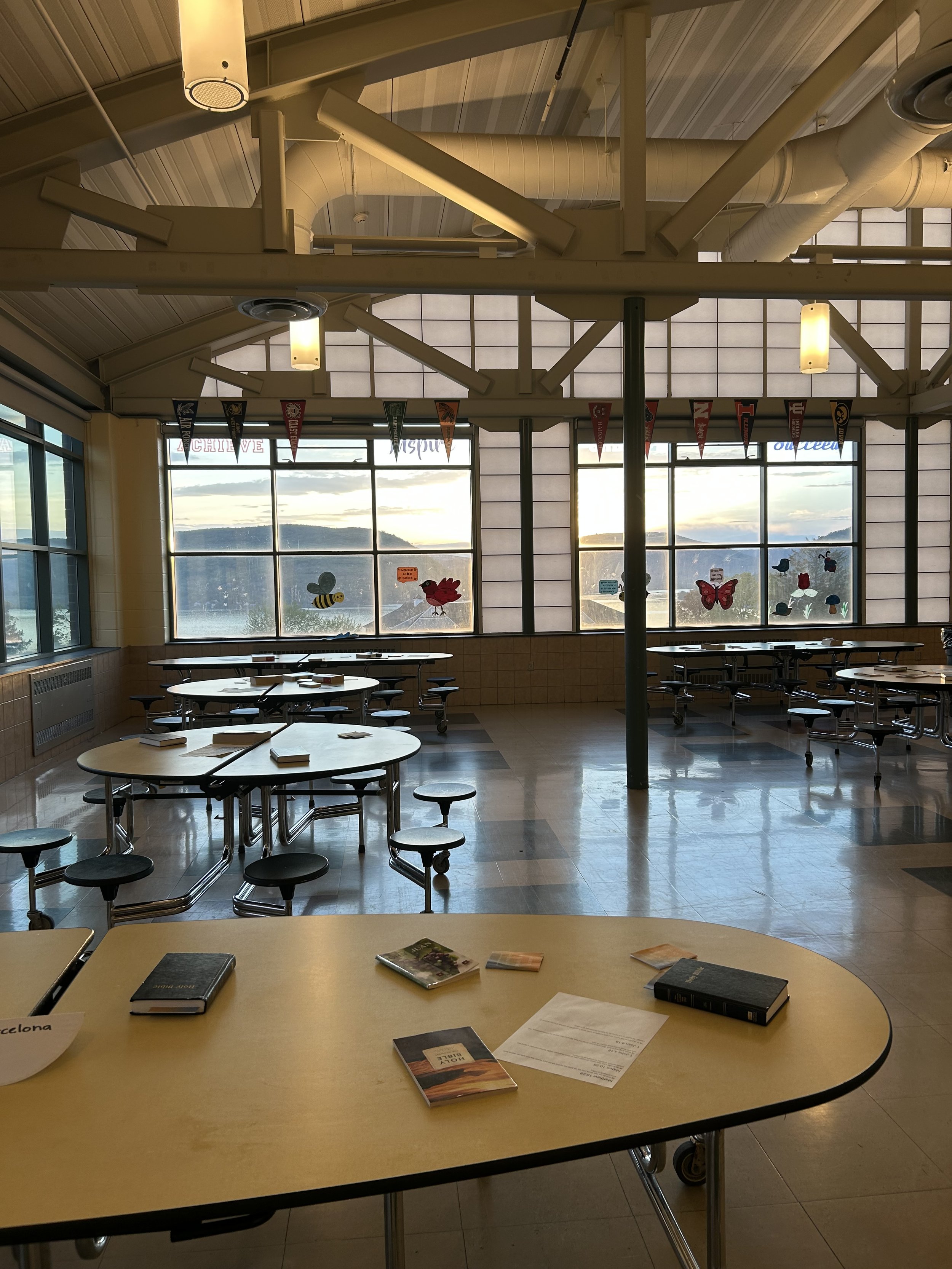 Empty school lunchroom with large windows showing a mountainous landscape at sunset, decorated with butterfly and bee stickers, and Bibles and tracts on the tables.