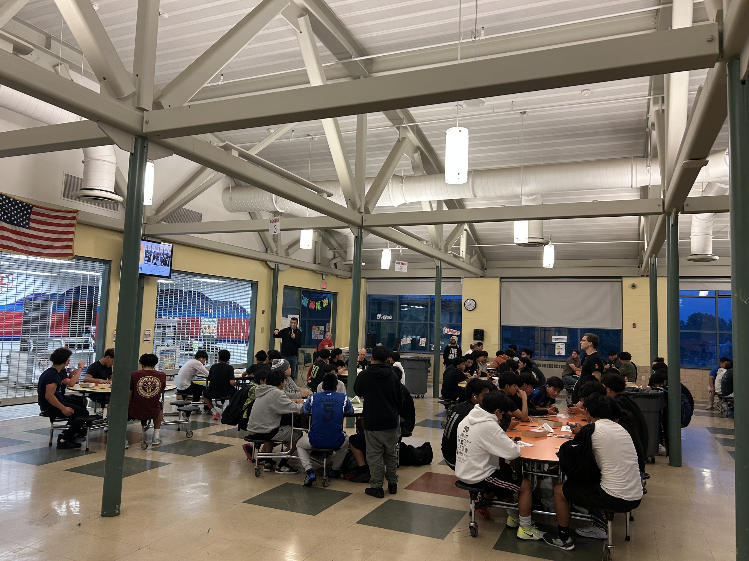 Group of students sitting at tables and listening to a speaker in a school cafeteria with large windows.