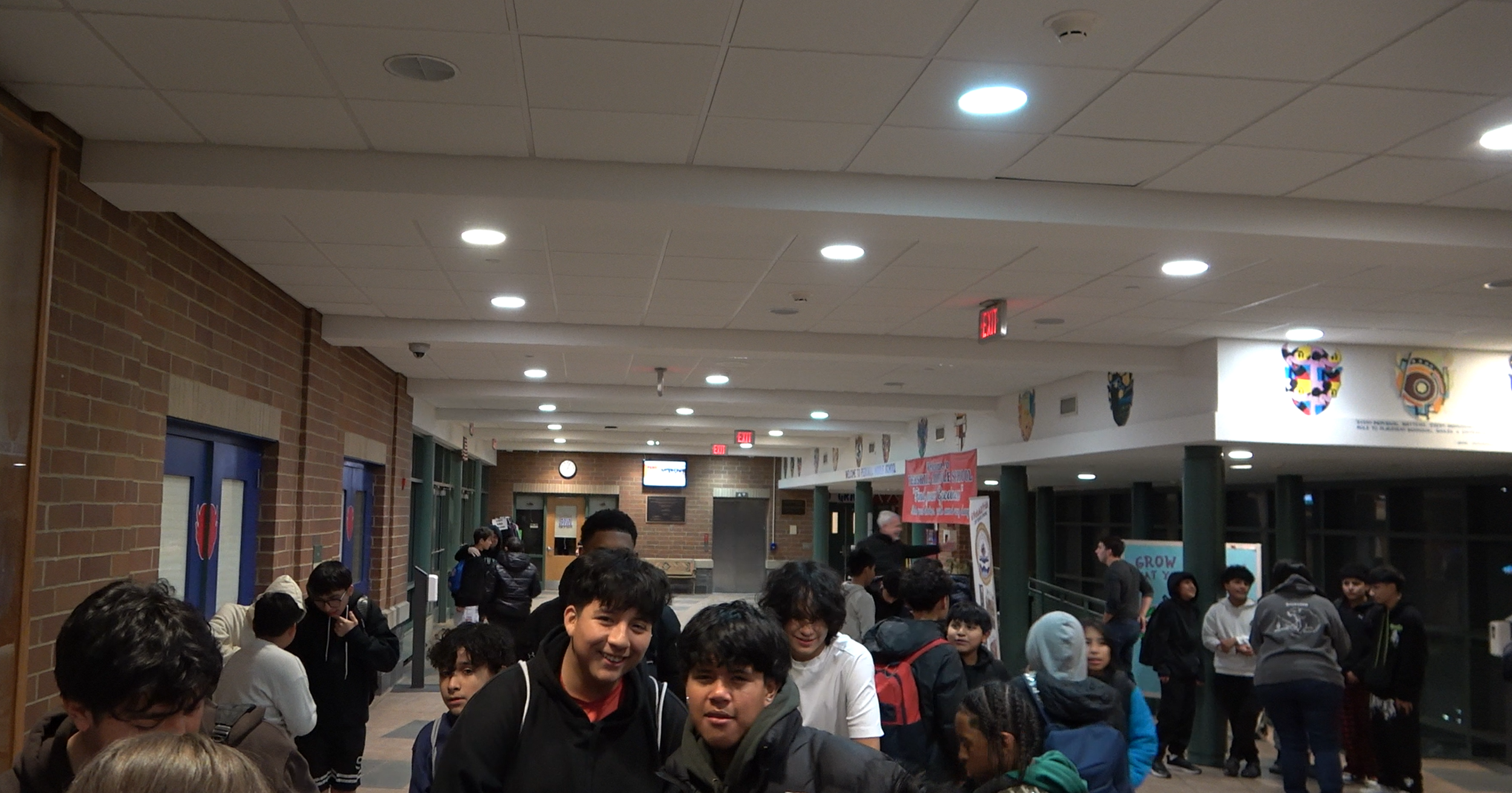 A group of students gathered in a school hallway with brick walls and display banners, some talking and others waiting.