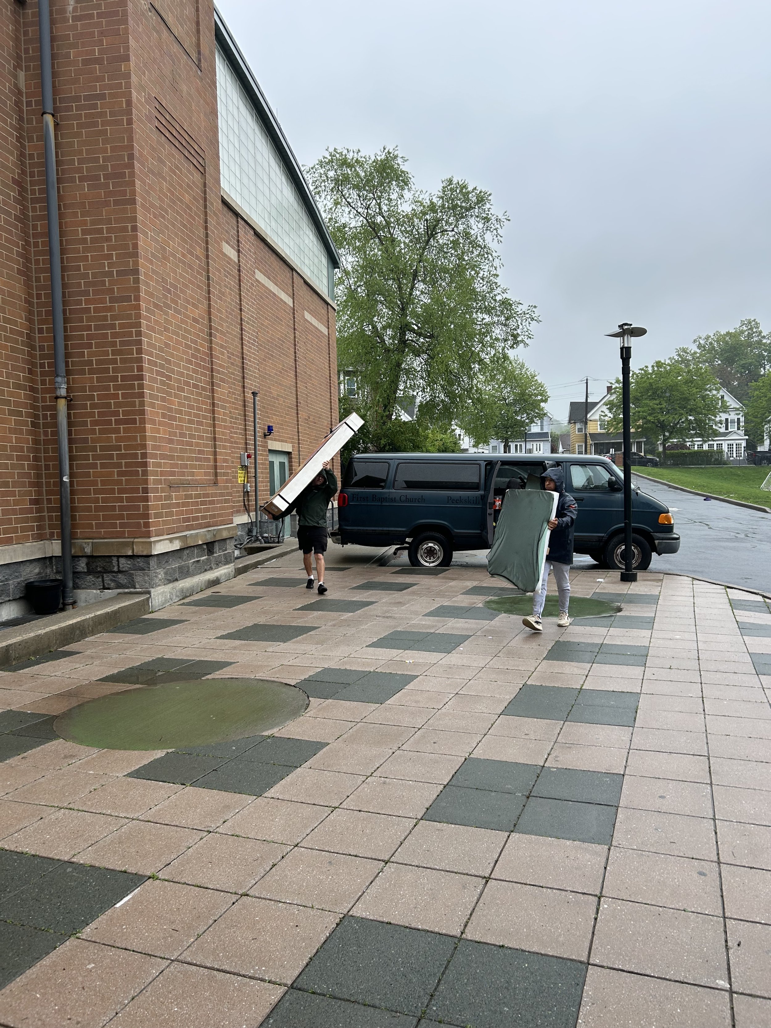 Two people are carrying large objects towards a van parked outside a brick building on a rainy day.