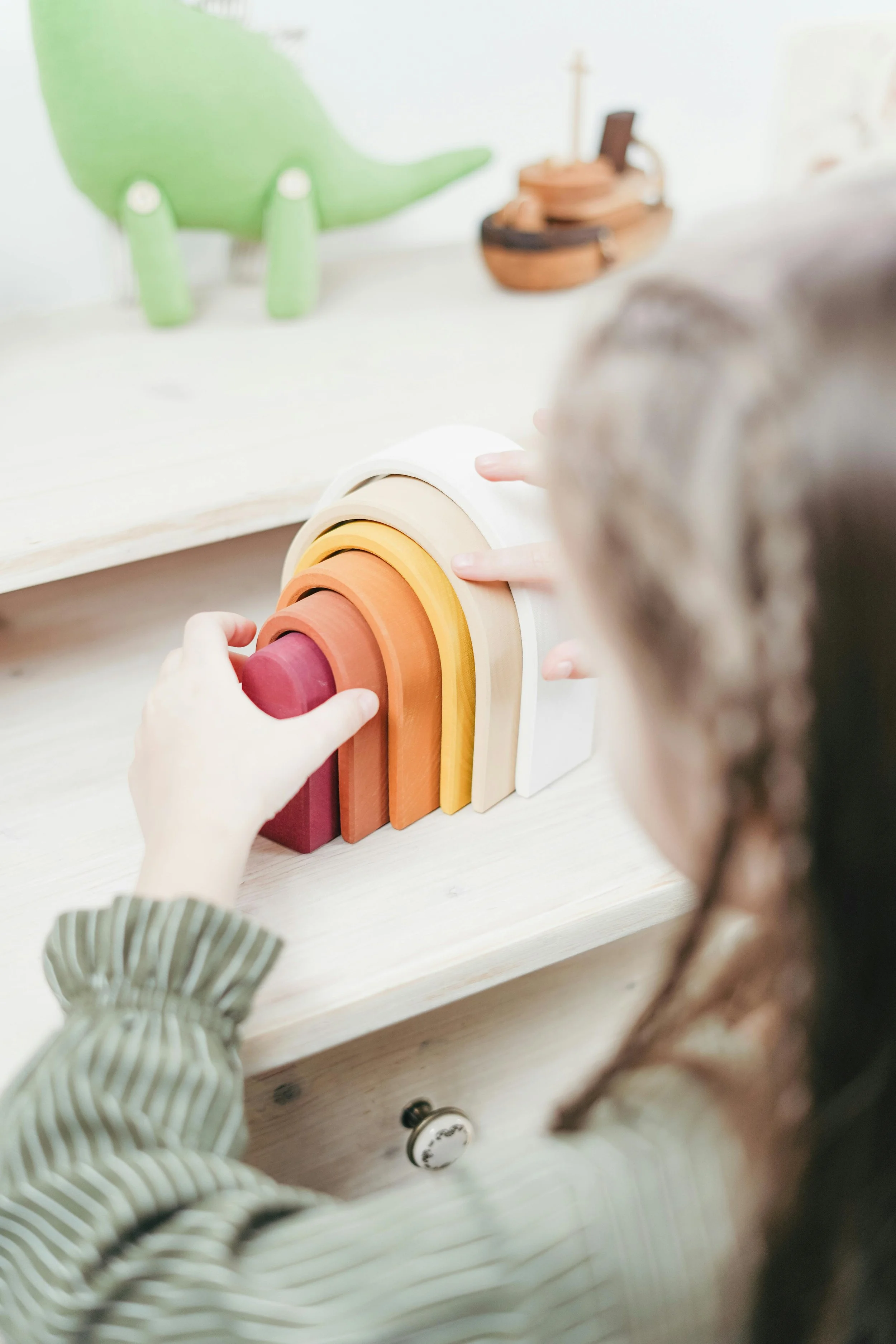 Child playing with a rainbow stacking toy in pastel colors on a light wooden surface.