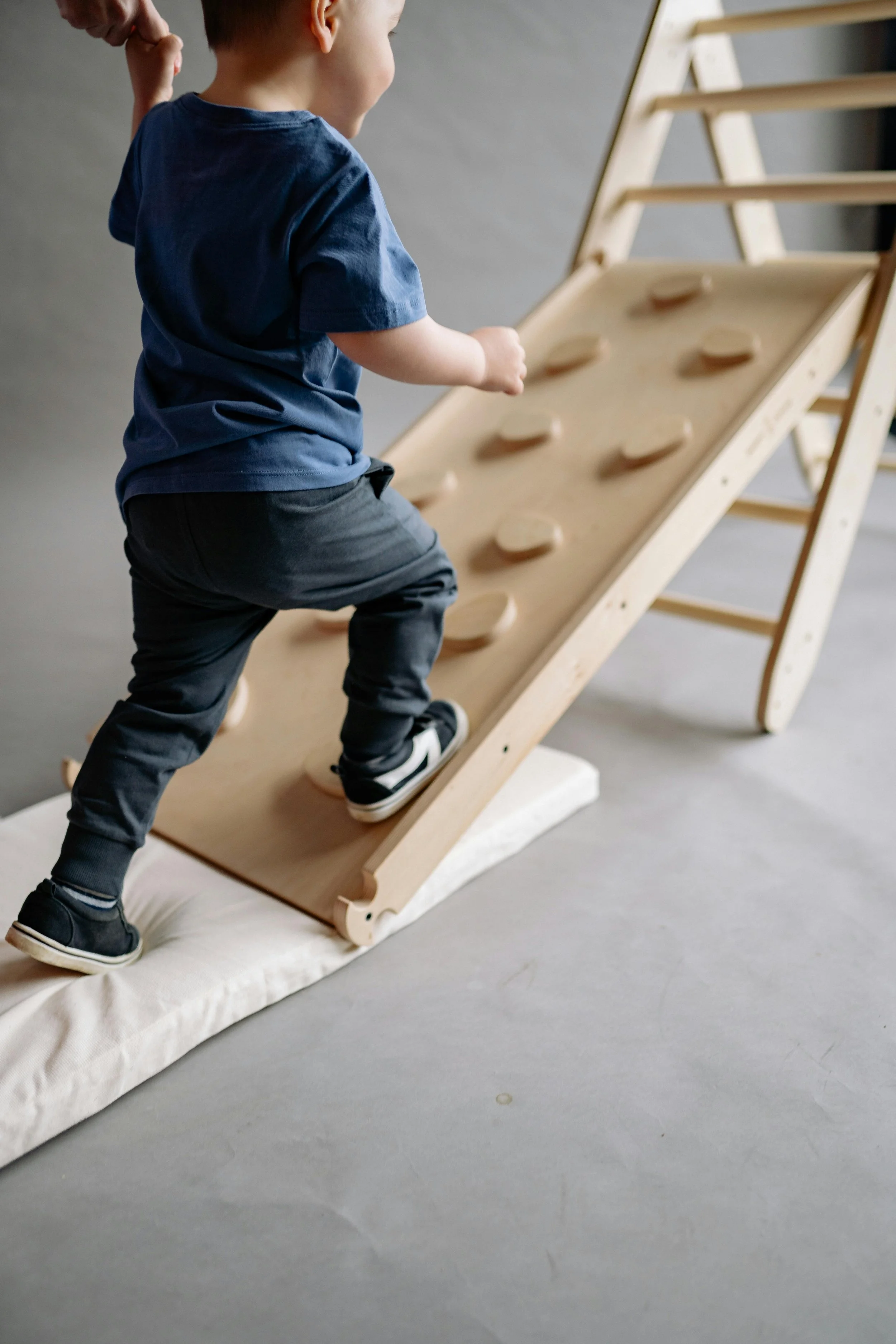 A young boy in a blue t-shirt and dark pants climbing a wooden indoor slide with a mat underneath.