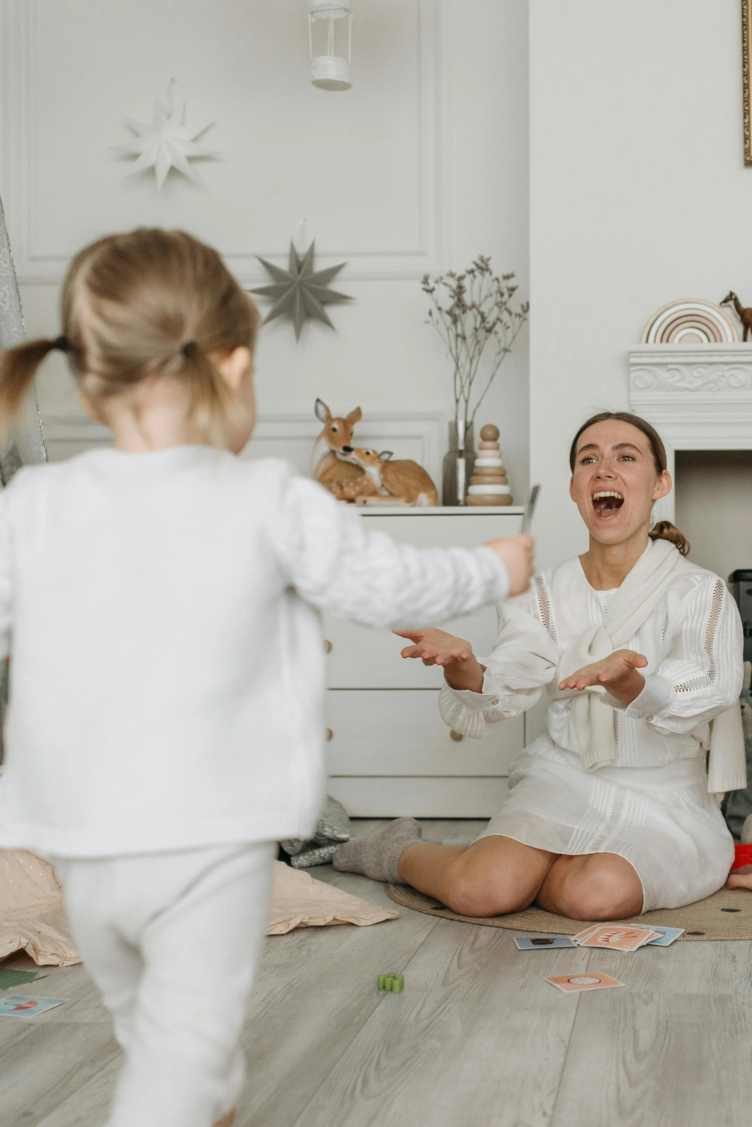 A woman sitting on the floor with her hands raised, playing a game with a young girl who is reaching out to her, in a cozy decorated room with toys and holiday decorations.