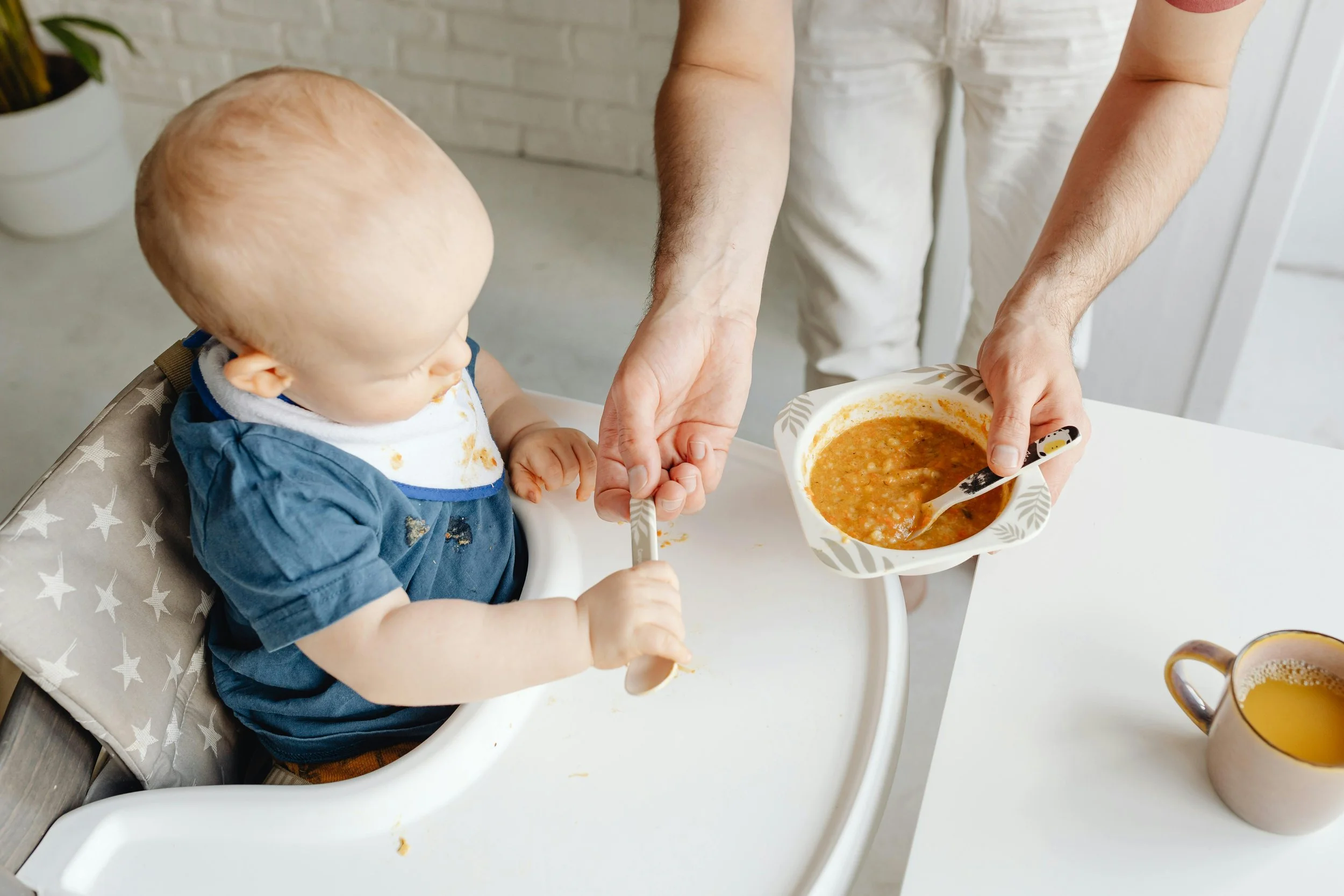 A baby sitting in a high chair being fed soup by an adult. The baby has a messy face and is holding a spoon. The adult is holding a bowl of soup and spoon, helping feed the baby. There is a cup of orange juice on the table.