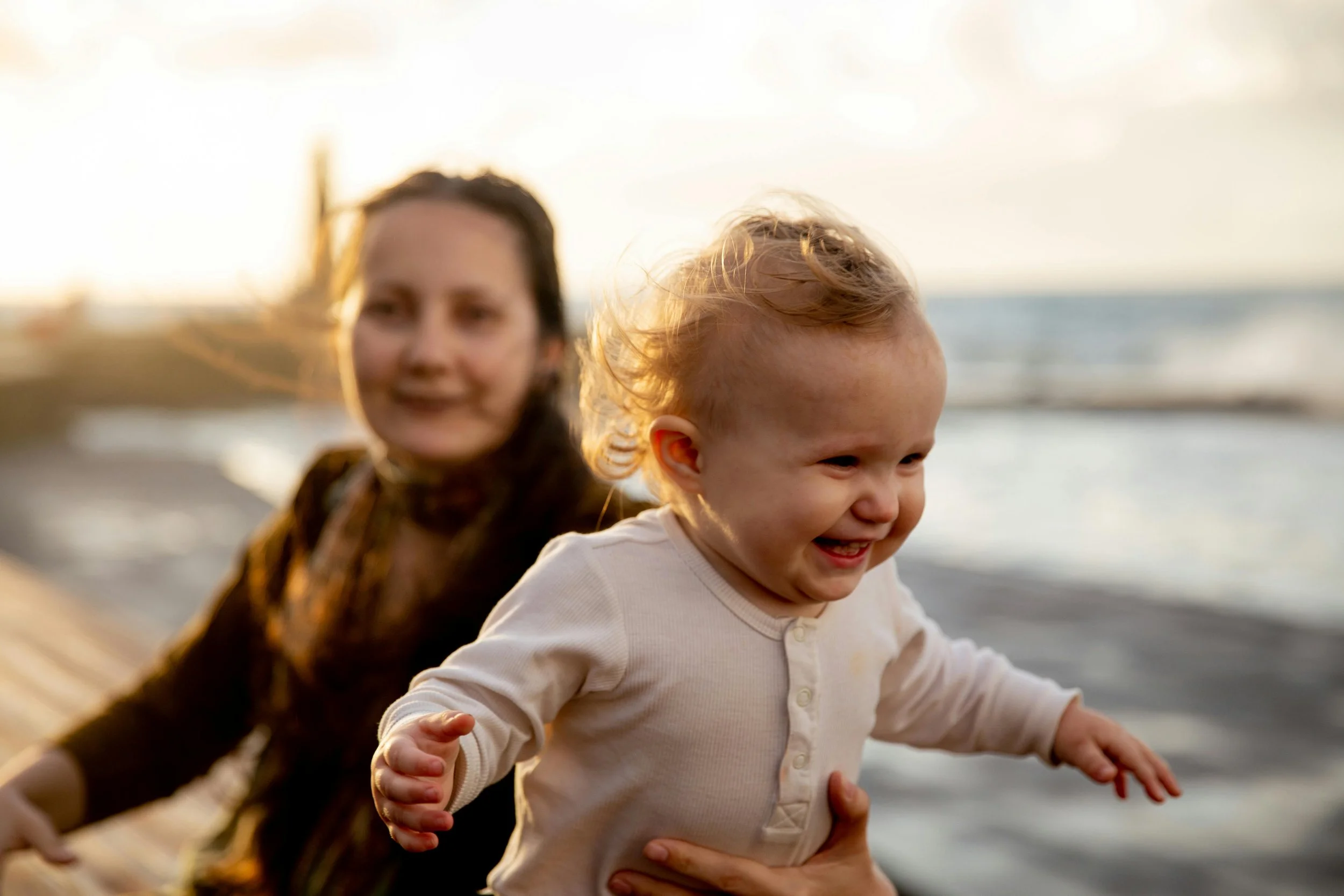 A woman with dark hair smiling in the background while a joyful toddler with curly hair is in a cream-colored shirt, being held and running near the beach at sunset.