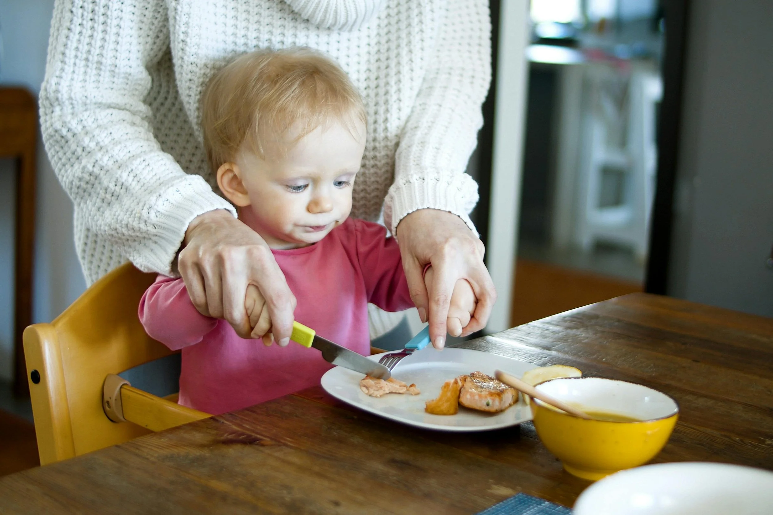 A young child sitting in a high chair at a wooden table, with an adult standing behind, helping them cut food on a plate.