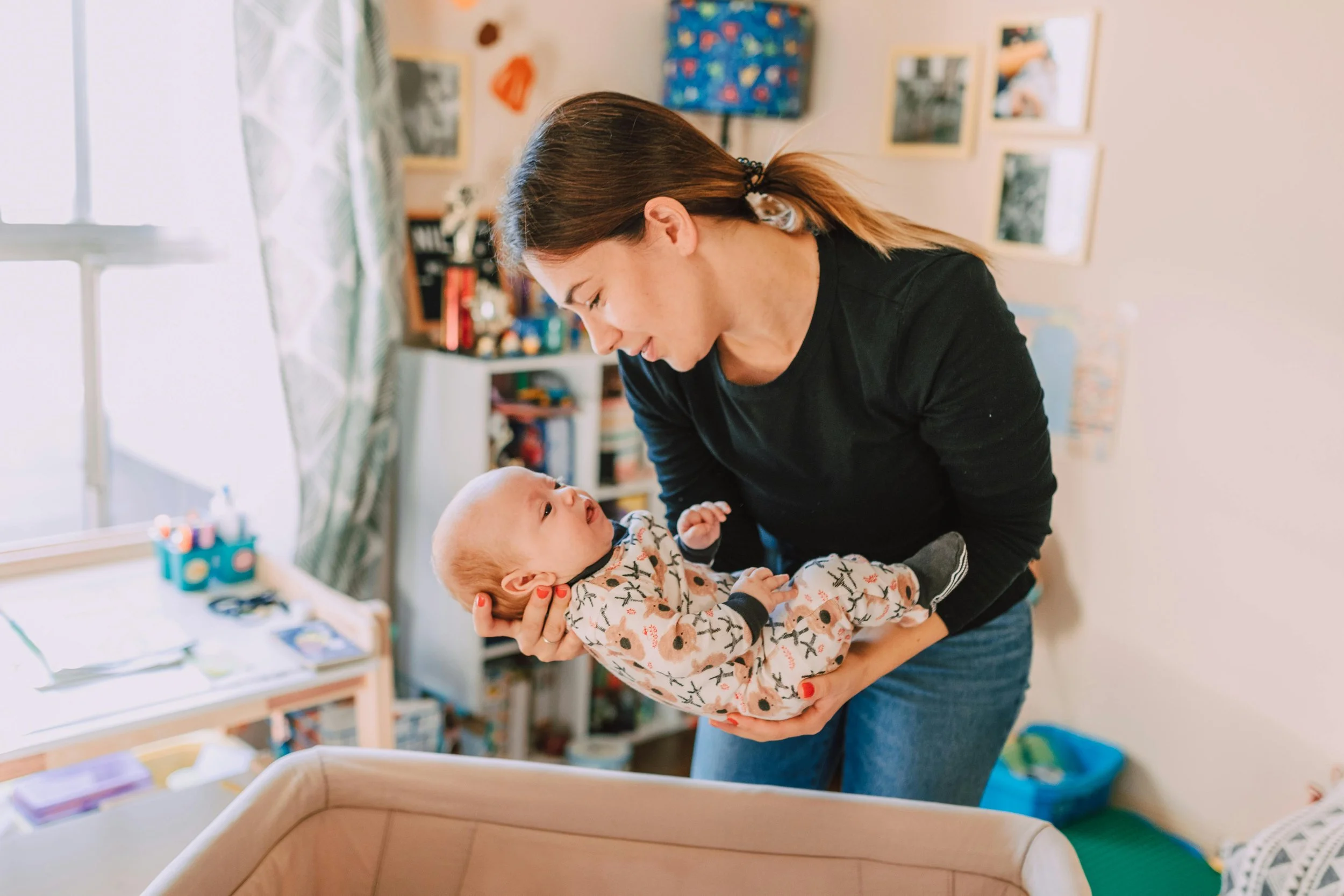 A woman holding a baby inside a home, looking at each other with smiles. The room has a window, shelves, and colorful decorations.