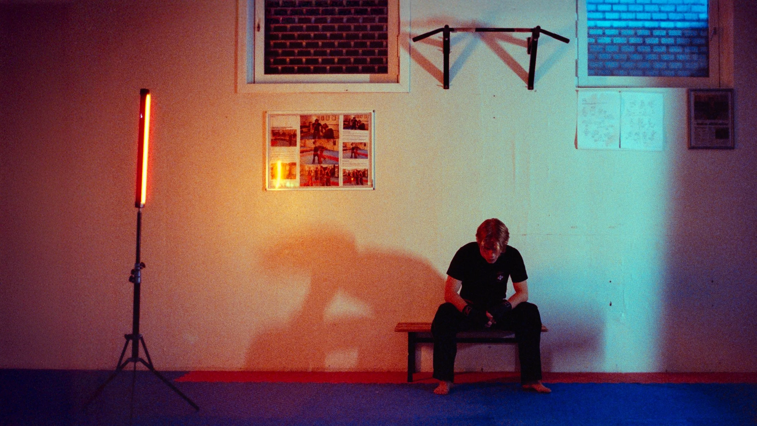 A person sitting alone on a bench in a room with yellow walls, illuminated by red and blue lighting, with a large red light on a stand to the left. The person appears to be looking down and is wearing black clothes.