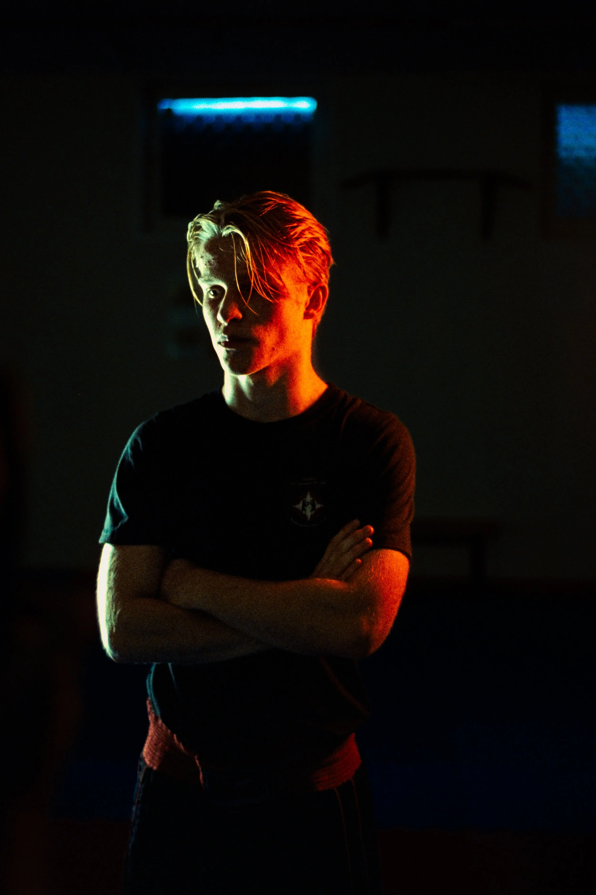A young man with blonde hair stands with his arms crossed in a dimly lit room, illuminated by red and yellow lights.