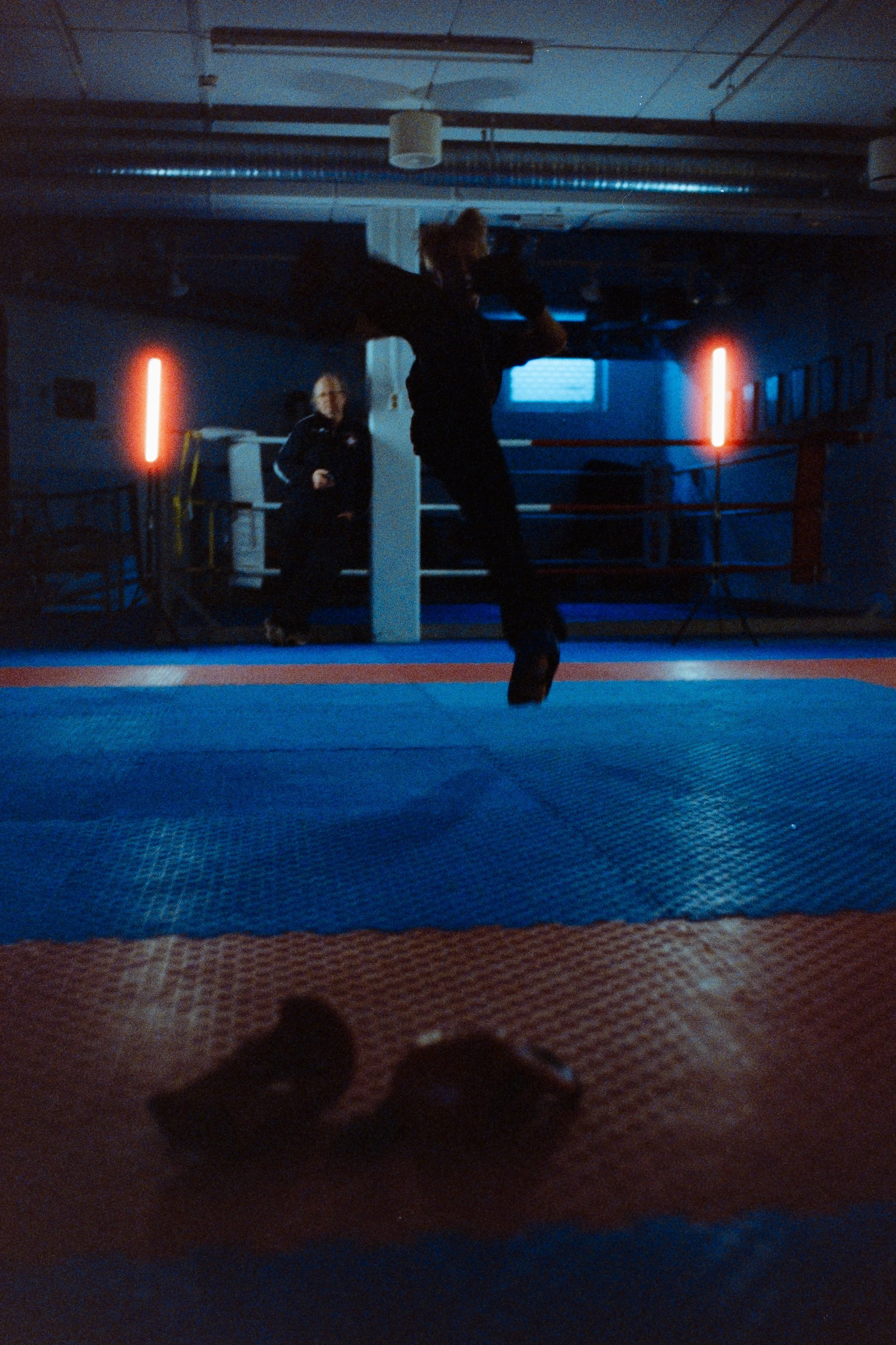 A person practicing martial arts or kickboxing in a dimly lit gym, with another person observing in the background. The gym has a boxing ring, and the floor has blue and orange mats. There are red LED lights on the walls.
