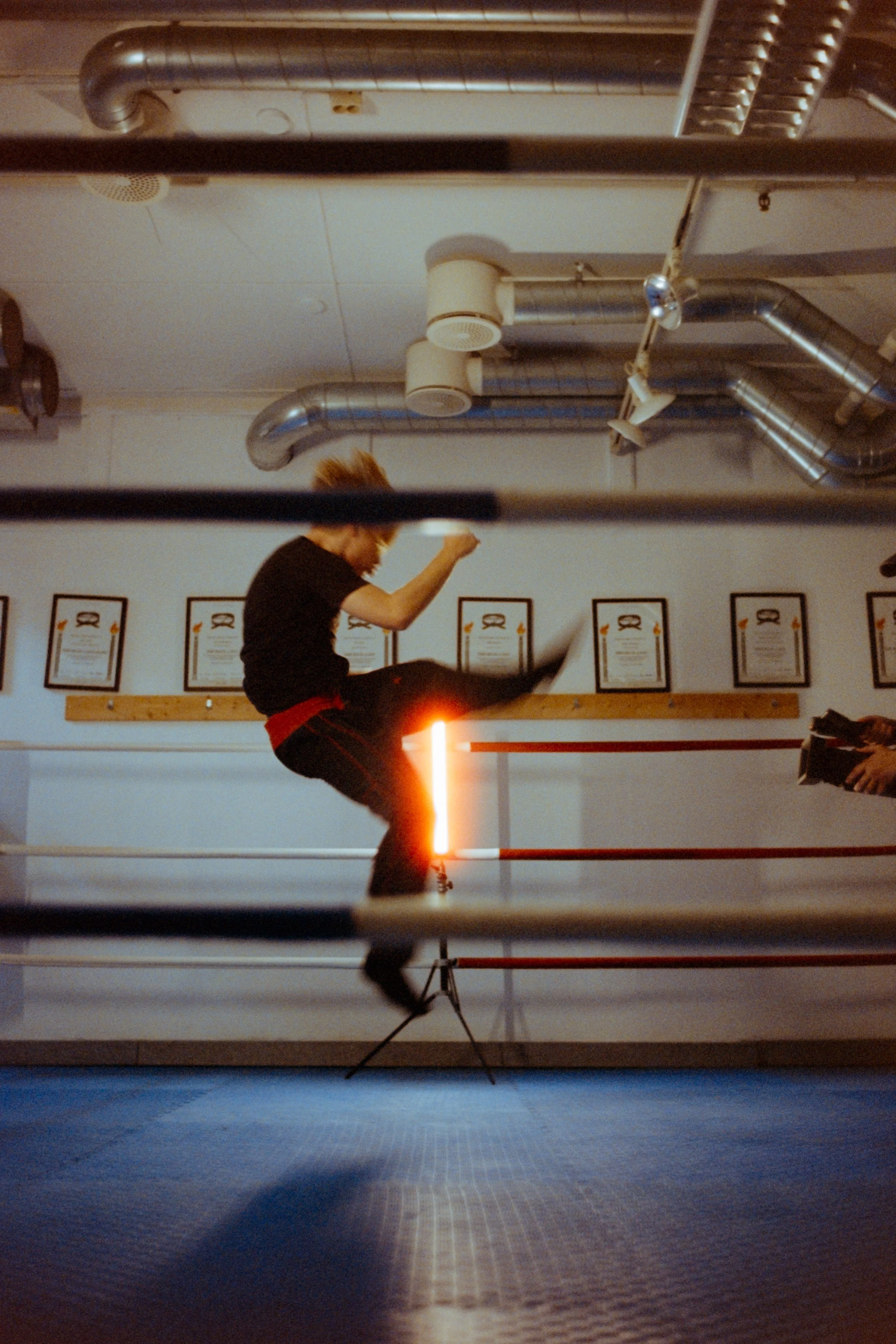 A person practicing kickboxing or martial arts in a gym, captured mid-air with a kick, with framed certificates or awards on the wall behind them.