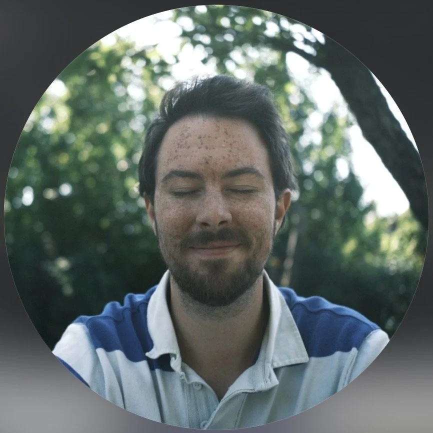 A man with a beard and closed eyes outdoors on a sunny day, wearing a white and blue polo shirt, with a background of trees and sunlight filtering through leaves.