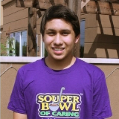 Young man wearing a purple T-shirt standing outdoors with a background of a building with windows and wooden panels.
