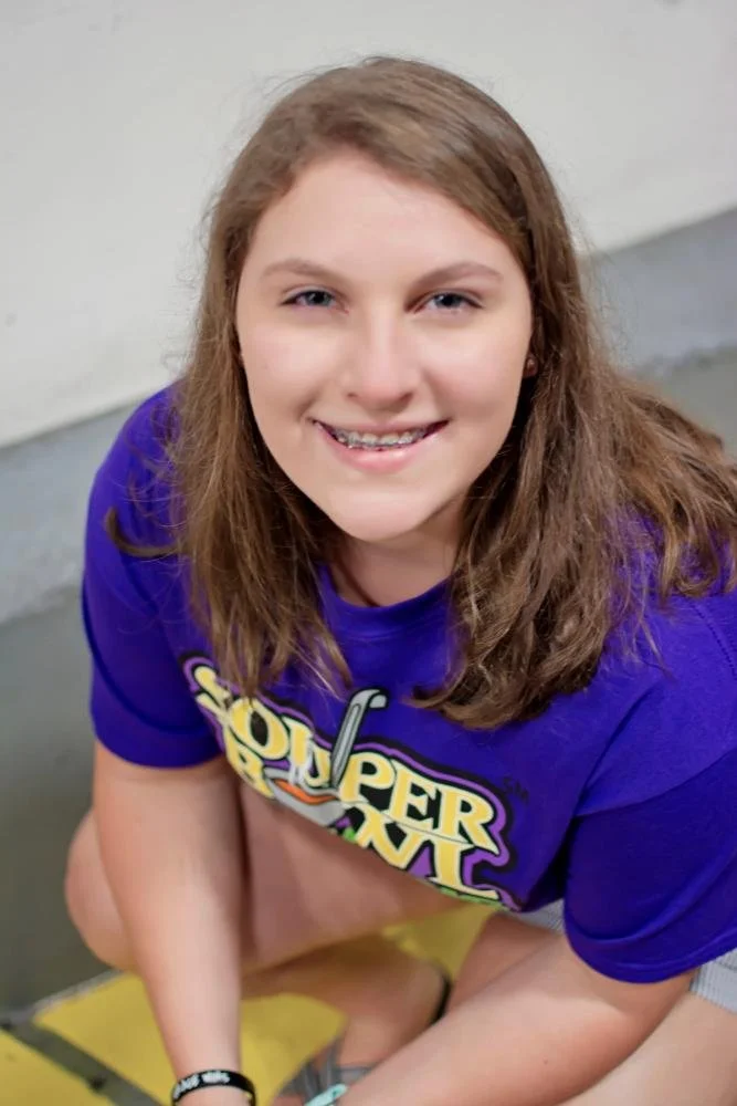 A smiling girl with braces, wearing a purple Super Mario T-shirt, sitting on a yellow and black floor mat.