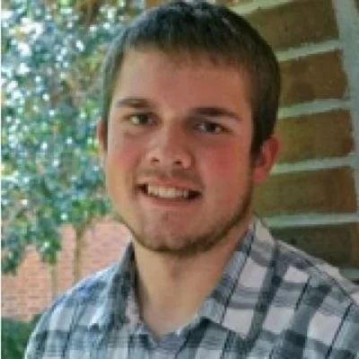 Young man with short brown hair, wearing a plaid shirt, standing outdoors near a brick wall, smiling.