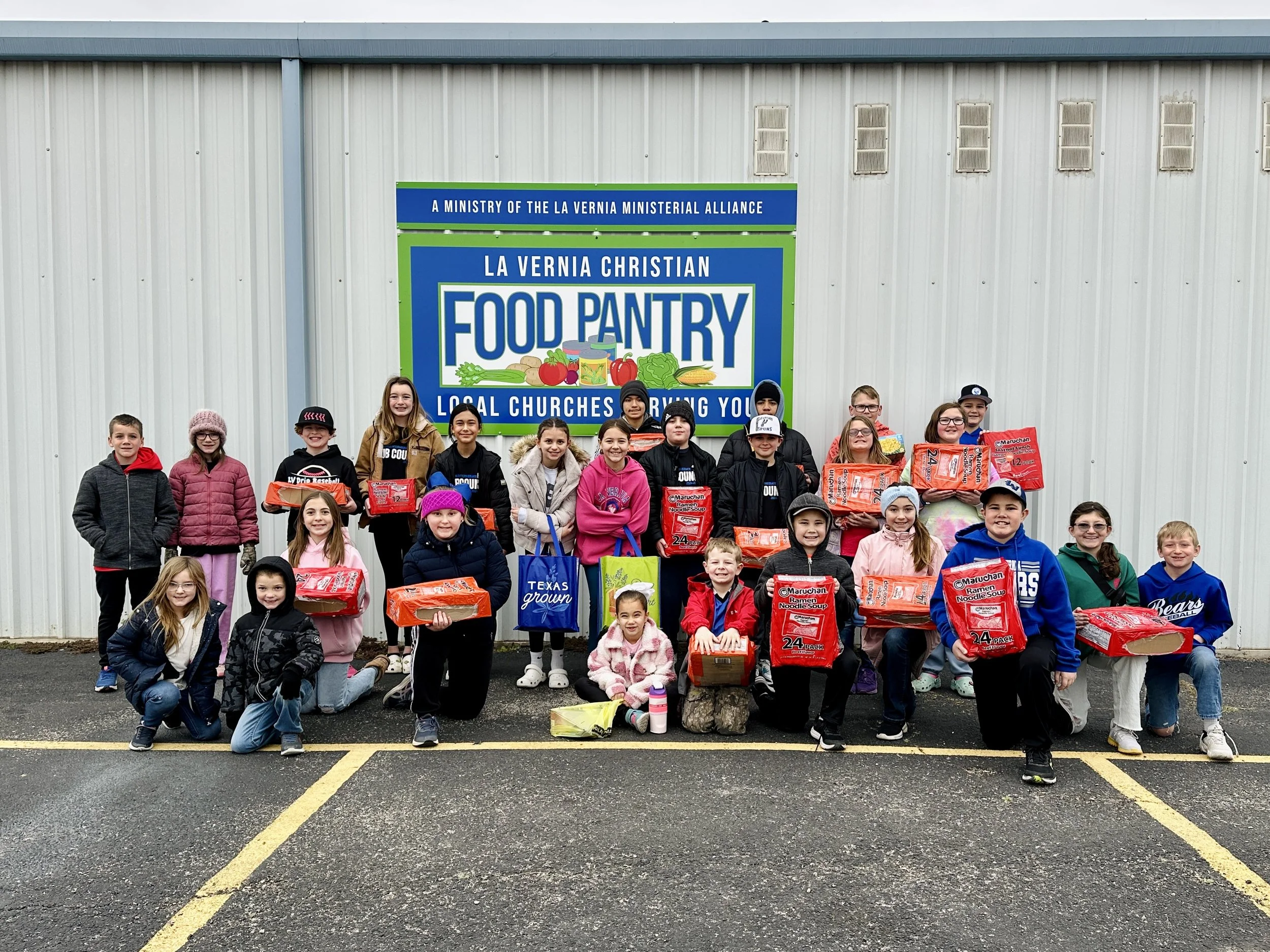 Group of children holding food donations in front of a large food pantry sign outside a building.