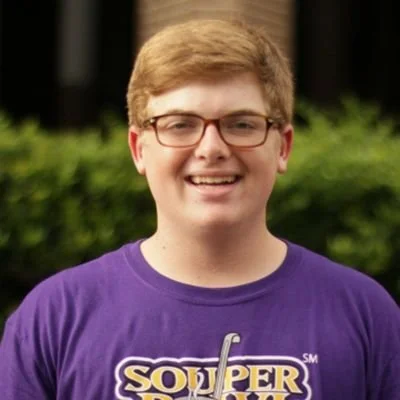 A young man with short, light brown hair and glasses smiling outdoors, wearing a purple T-shirt with yellow and white text.