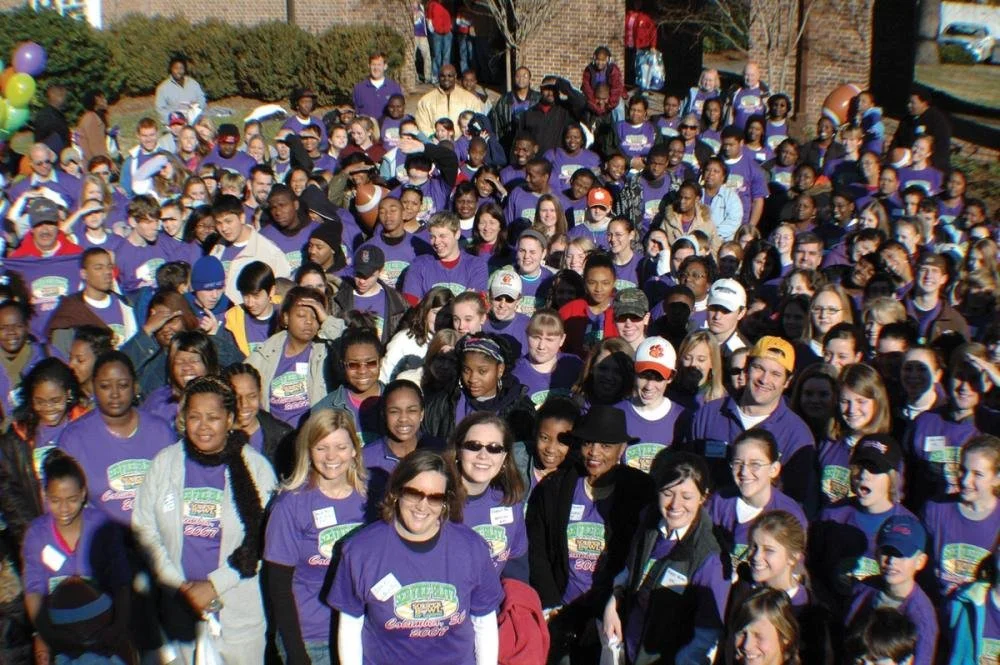A large group of children and adults gathered outdoors for a community event, wearing purple T-shirts.