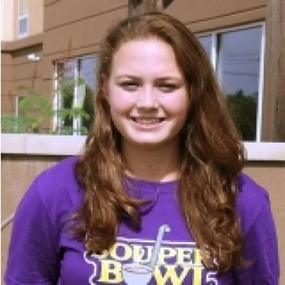 Young woman with long, curly hair smiling outdoors in front of a building, wearing a purple T-shirt.