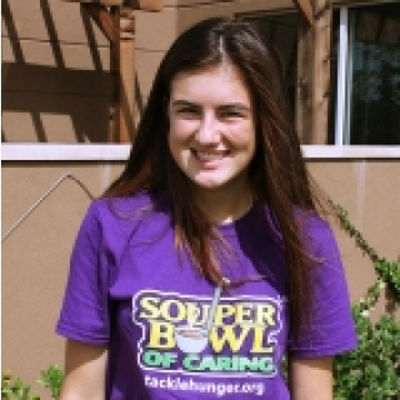 Smiling young woman with brown hair wearing a purple shirt that says 'SOUper BOWL OF CARING' and standing outdoors near a house with greenery.