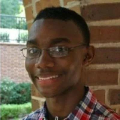 Young man with glasses, smiling, wearing a plaid shirt, standing outside near a brick wall.