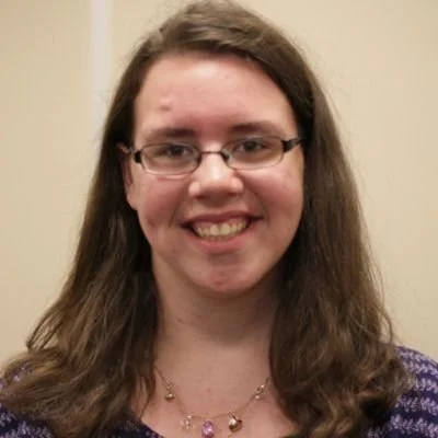 A woman with long brown hair, glasses, and a purple patterned top, smiling at the camera.