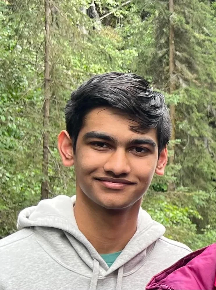 Young man with dark hair and a light gray hoodie standing outdoors in a forested area with green trees in the background.