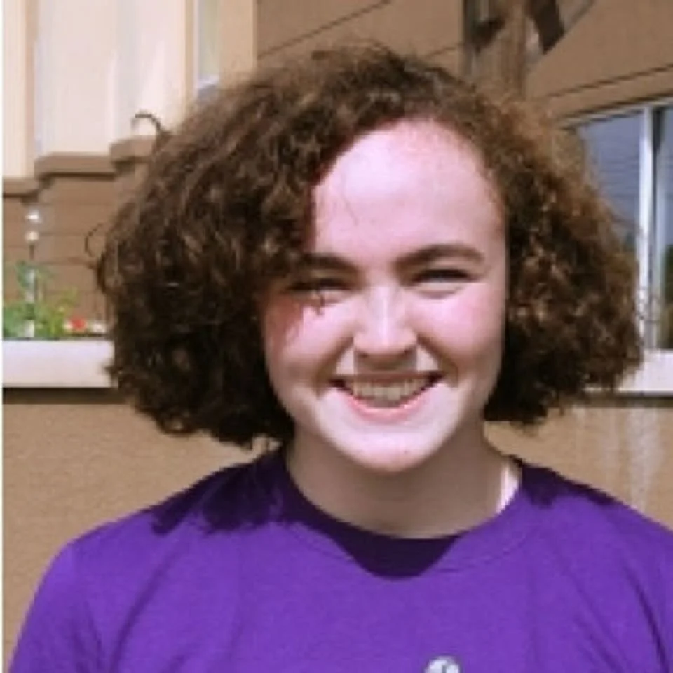 A young woman with short, curly brown hair smiling outdoors in front of a house.