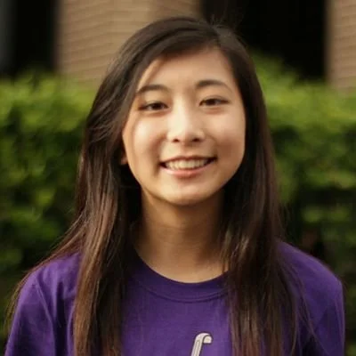 Young woman with long dark hair smiling outdoors, wearing a purple shirt.