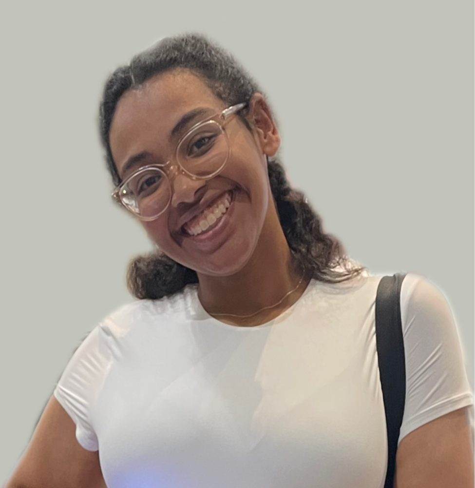 A smiling woman with glasses and curly hair, wearing a white shirt and carrying a black strap, standing against a plain background.