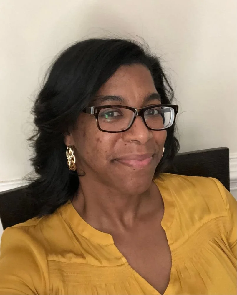 A woman with black hair, glasses, and earrings, wearing a yellow top, sitting on a chair with a neutral background.