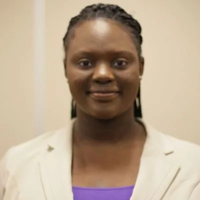 A woman with braided hair, wearing a light-colored blazer over a purple top, looking at the camera with a neutral expression.