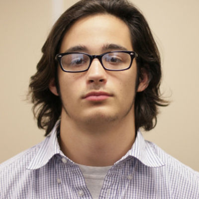 A young man with long brown hair, glasses, and a striped shirt looking down thoughtfully.