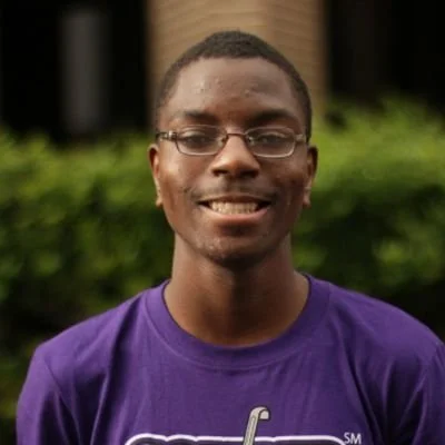 Young man with glasses smiling, wearing a purple shirt, outdoors with greenery in the background.