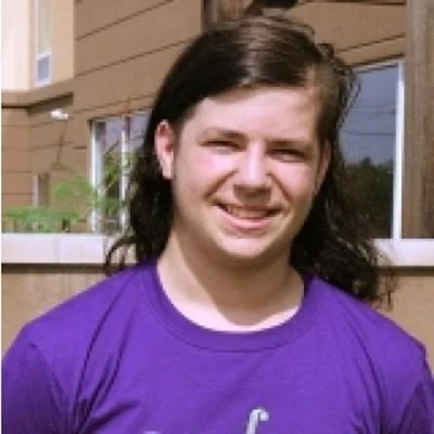 A young woman with dark brown hair wearing a purple shirt, standing outdoors in front of a brown house with windows.