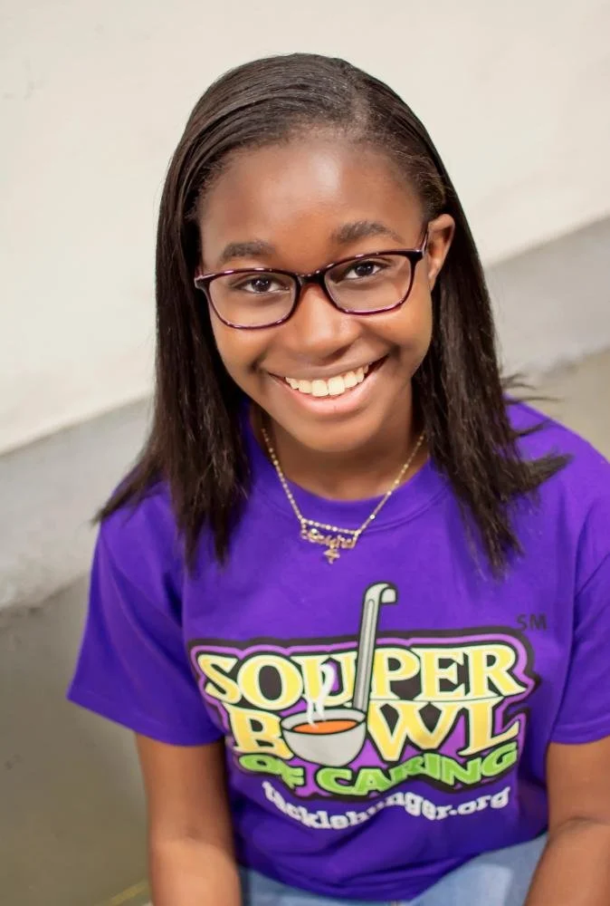 Smiling young girl with glasses wearing a purple T-shirt promoting the Souper Bowl of Caring fundraiser.