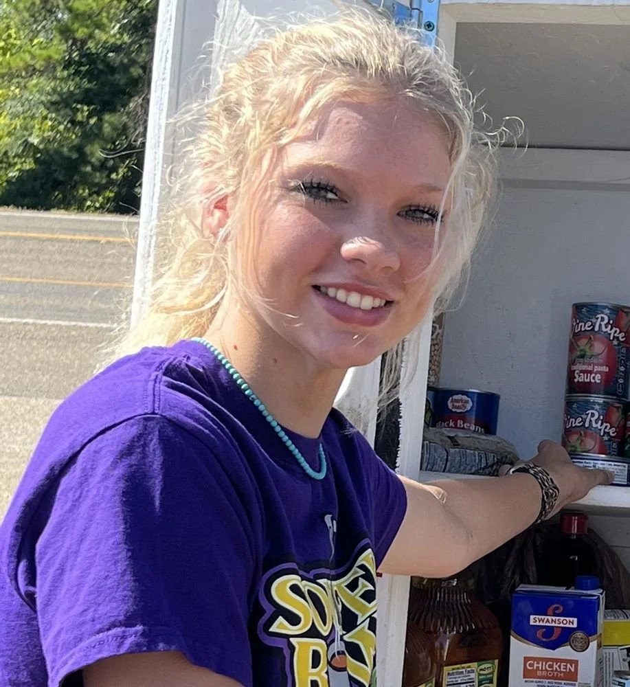 A young woman with blonde hair wearing a purple shirt and a beaded necklace, smiling and reaching into a grocery cart filled with canned goods and food items, outdoors on a sunny day.