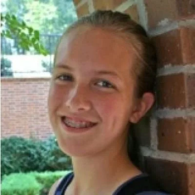 A young girl smiling outdoors, leaning against a brick wall with greenery and a fence in the background.