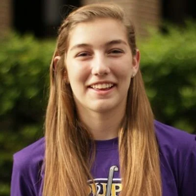 A young woman with long blonde hair smiling outdoors, wearing a purple shirt.