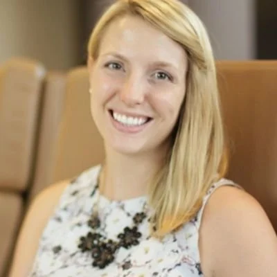 A woman with blonde hair smiling while sitting on a brown chair in an indoor setting.