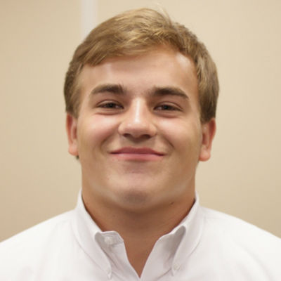 Close-up image of a young man with short blonde hair, wearing a white collared shirt, smiling at the camera.