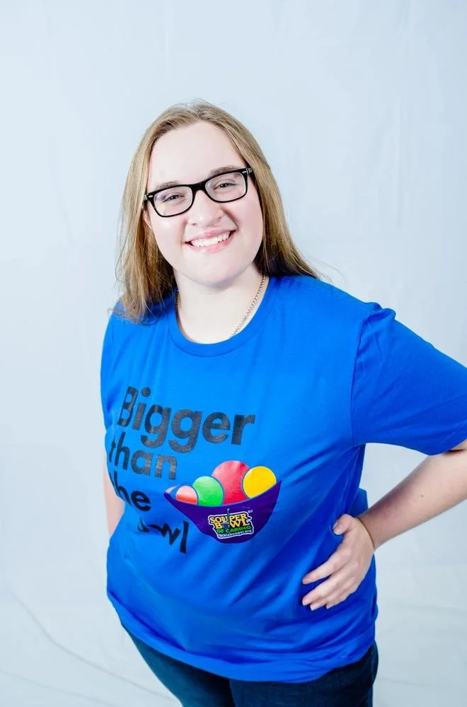 Young woman with glasses and long hair wearing a blue T-shirt with the text 'Bigger than the' and a colorful bowl of candies graphic.