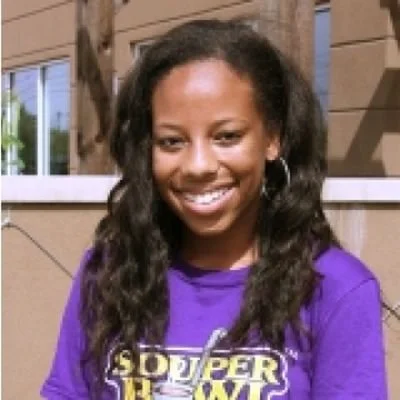 Young girl with curly hair smiling, wearing a purple shirt with a design, standing outdoors near a wooden fence.